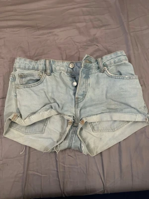 Ljusblå denimshorts med rå kant - Säljer ett par ljusblå denimshorts med hög midja och råa, fransiga kanter. Shortsen har klassisk femficksdesign, knappgylf och bälteshällor. Perfekta för sommaren och ger en avslappnad vibe.