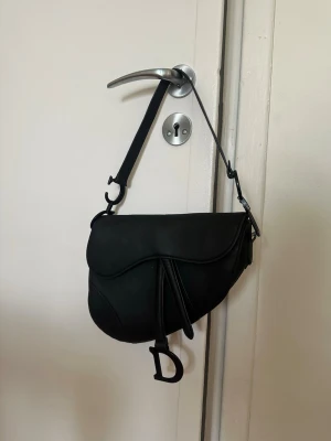 Svart handväska - Säljer en svart saddle handväska i läderimitation med unik form och en stor D-detalj framtill. Väskan har ett kort axelband och stilrena sömmar som ger en modern look. Perfekt accessoar för att lyfta din outfit.