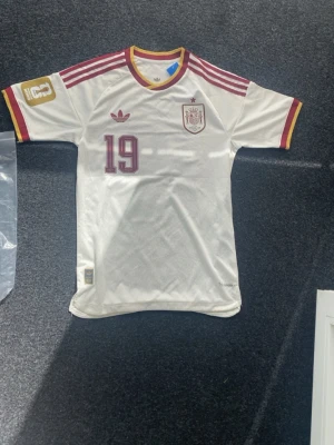 Spaniens bortaställ 2026  - Säljer en vit Spanien fotbollströja från Adidas med vinröda detaljer och nummer 19. Tröjan har Lamine Yamal tryckt på ryggen, röd-gula ränder på axlarna och klubbmärke på bröstet. Perfekt för dig som älskar fotboll och vill stötta landslaget.