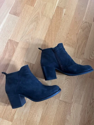 Svarta stövletter med klack - Snygga svarta stövletter i mockaimitation med bred klack och dragkedja på sidan. Skorna har rundad tå och en enkel, stilren design som passar till många olika outfits. Perfekta för dig som gillar en klassisk look med lite edge.