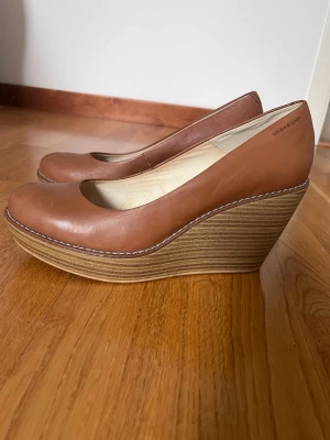 Bruna kilklackade pumps från Vagabond - Snygga bruna pumps från Vagabond med rund tå och hög kilklack i trälook. Skorna är i slätt läder och har vita sömmar längs kanten för en clean och stilren look. Perfekta för dig som vill ha både höjd och komfort.