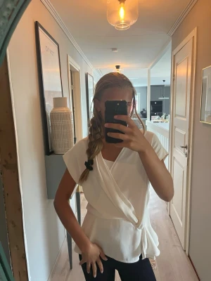Vit blus - Snygg vit omlottopp med knytband i midjan och korta ärmar. Toppen har en avslappnad passform och ett mjukt, lätt tyg som ger en stilren look. Perfekt att matcha med jeans eller kjol för en trendig outfit.