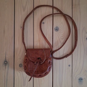 Boho Vintage Läderhandväska - Handväska i äkta läder med långt axelband. Väskan har Peru och ett landskap graverat på framsidan och en fågel med ett landskap på baksidan. Djup ca 10 cm. Längd ca 12 cm. Höjd ca 12,5 cm. Bredd ca 5 cm. Axelrem längd ca 120,5 cm. Kan ha mindre små märken av slitage.