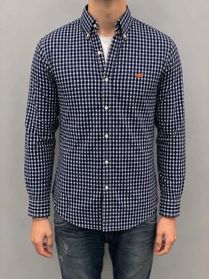 Hollister skjorta - Snygg mörkblå och vit rutig skjorta med lång ärm och knappkrage. Skjortan har vita knappar framtill och en liten orange logga broderad på bröstet. Tillverkad i mjukt bomullstyg och passar perfekt till jeans för en avslappnad look.