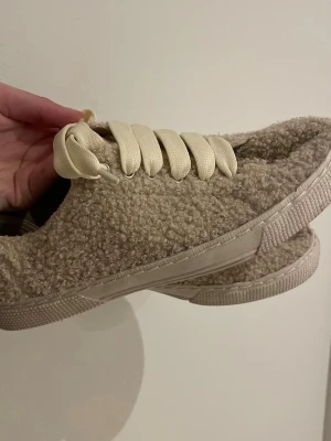 Beige teddy-sneakers med breda snören - Unika sneakers i beige teddy-material med breda, krämvita snören och matchande sula. Skorna har en låg profil och en chunky känsla som sticker ut. Perfekta för dig som vill ha något annorlunda och mysigt till din outfit.