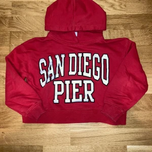 Hoodie från Zara - Säljer en använd röd hoodie från Zara med stor vit text 'San Diego Pier' på bröstet. Tröjan har huva. Tröjan är för tjejer och halva magen syns pga modellen. Tröjan är använd ett par gånger, men har inga tydliga tecken på användning förutom att den tyvärr har en namnlapp vid nacken. 