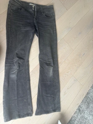 Grå bootcut jeans med fickdetaljer - Säljer ett par grå bootcut jeans med klassisk femficksmodell och snygga ficklock med knappar bak. Jeansen har normal midja och subtila slitningar framtill för en avslappnad look. Tillverkade i mjukt denimtyg med lite stretch för skön passform. Dom e väldigt använda men inga slitningar vid fötterna 