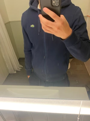 Mörkblå hoodie från Lacoste - Snygg mörkblå hoodie från Lacoste med dragkedja framtill och klassisk krokodillogga på bröstet. Modellen har huva med dragsko och ribbade muddar vid ärmslut och nederkant. Perfekt för en chill och sportig stil.