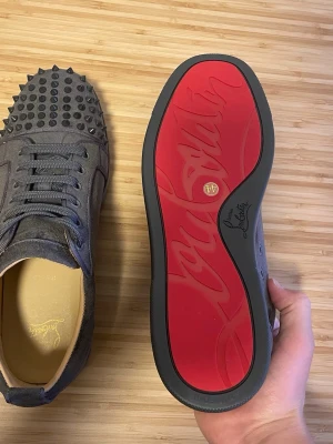 Gråa sneakers med nitar och röd sula - Snygga gråa sneakers i mocka från Christian Louboutin med ikonisk röd sula och svarta nitar på tån. Skorna har rundad tå, matchande grå skosnören och en lyxig känsla. Perfekta för dig som vill sticka ut med en edgy och exklusiv stil.