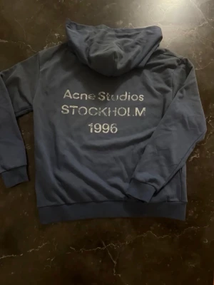 Blå hoodie från Acne Studios M - Mörkblå hoodie från Acne Studios med tryck på ryggen: 'Acne Studios STOCKHOLM 1996'. Klassisk passform med huva och ribbade muddar. Perfekt för en chill och stilren look.