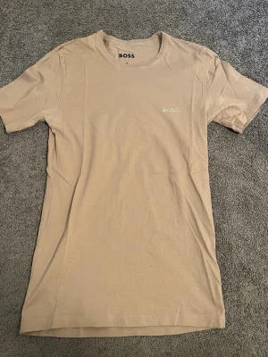 Beige t-shirt från BOSS - Snygg beige t-shirt från BOSS med diskret broderad logga på bröstet. Klassisk rund halsringning och korta ärmar. Perfekt för en clean och stilren look.