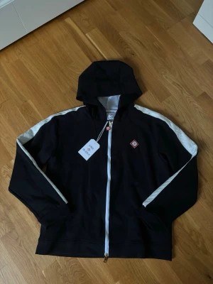 Casablanca zip-up Hoodie  - Snygg svart hoodie från Casablanca med vit dragkedja och vita detaljer längs ärmarna. Märkets logga i rött och vitt sitter på bröstet och dragkedjan har en matchande logga. Hoodien har huva och är tillverkad i mjukt bomullsmaterial.
