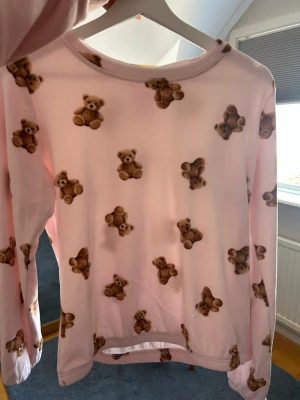 Rosa loungewear-tröja med nallemönster - Supersöt långärmad loungewear-tröja i ljusrosa med gulligt nallebjörnsmönster över hela plagget. Mjuk och bekväm, perfekt för mysiga hemmakvällar. Rund halsringning och avslappnad passform. Materialet känns lent och stretchigt.