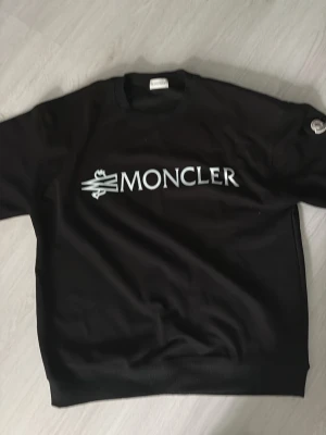 Svart sweatshirt från Moncler - Snygg svart sweatshirt från Moncler med stor vit logga framtill. Tröjan har rund halsringning och ribbade muddar vid ärmslut och nederkant. Perfekt för en clean och stilren look. Start pris 800kr