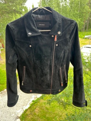 Svart skinnjacka från Vero Moda - Snygg svart skinnjacka från Vero Moda med asymmetrisk dragkedja, bred krage och silverfärgade detaljer. Jackan har tre dragkedjeförsedda fickor framtill och en cool bikerstil. Perfekt för dig som vill ha en stilren och trendig look.