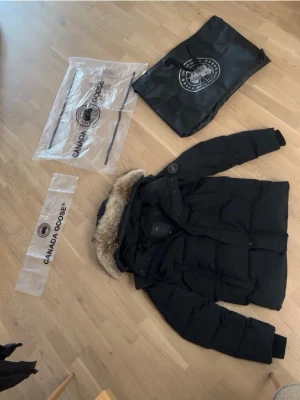 Svart dunjacka med päls från Canada Goose - Säljer en svart dunjacka från Canada Goose med huva och snygg pälsdetalj. Jackan har klassisk puffer-design, logotyp på ärmen och flera praktiska fickor. Perfekt för kalla vinterdagar och riktigt varm. Jackan levereras med originalpåse.