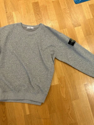 Grå sweatshirt från Stone Island  - Snygg grå sweatshirt från Stone Island med klassisk rund halsringning och ribbade muddar. Ikonisk svart patch med logga på vänster ärm ger en clean och trendig look. Perfekt till jeans eller cargo för en avslappnad stil.