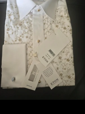 Vit skjorta med blommigt mönster Eton - Säljer en elegant vit skjorta från Eton med diskret blommigt mönster i beige toner. Skjortan har klassisk krage, långa ärmar och knappar framtill. Perfekt för dig som vill ha en stilren men unik look.