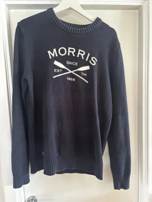 Mörkblå stickad tröja från Morris - Snygg mörkblå stickad tröja från Morris med vit logga och text framtill. Tröjan har rund halsringning, långa ärmar och ribbade muddar vid ärmslut och nederkant. 