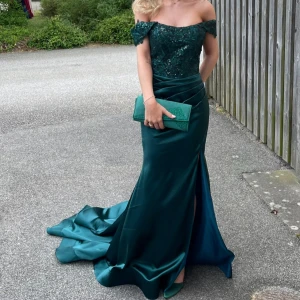 BALKLÄNNING - Elegant mörkgrön långklänning med offshoulder och vacker spets upptill. Klänningen har en figurnära passform, slits framtill och draperad midja. Perfekt för bal eller fest. Köpt för 1770kr på hebeos! Använd två gånger❤️