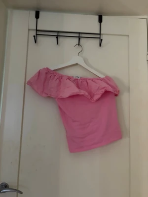 Rosa offshoulder topp med volang - Söt rosa offshoulder topp med bred volang runt axlarna. Toppen är ärmlös och har en croppad passform som ger en trendig look. Perfekt för varma dagar och somriga outfits.