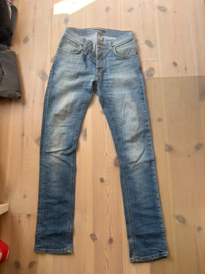 Blå raka jeans från Nudie Jeans - Säljer ett par klassiska blå jeans från Nudie Jeans med raka ben och snygg slitning. Jeansen har fem fickor, orange sömmar och Nudie-logga på bakfickan. Tillverkade i denim med en autentisk tvättad look. Storlek 30/32