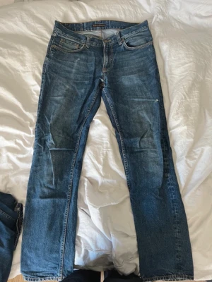 Nudie Jeans gritty jackson - Säljer ett par klassiska blå jeans från Nudie Jeans med rak passform och fem fickor. Jeansen har snygg slitning och kontrastsömmar samt Nudie-logga på bakfickan. Tillverkade i denim, perfekt för en avslappnad look.