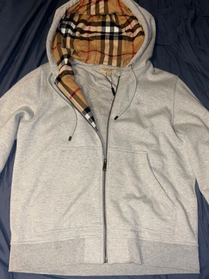 Grå zip hoodie från burberry - Säljer en ljusgrå hoodie från Burberry med dragkedja och klassiskt rutigt foder i huvan. Hoodien har dragsko i huvan, två fickor framtill och diskret Burberry-logga broderad på bröstet. Perfekt för dig som gillar stilrena plagg med exklusiva detaljer.