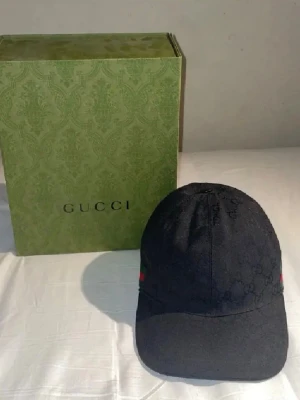 Svart keps från Gucci med monogram - Snygg svart keps från Gucci med klassiskt GG-monogram i tyget och diskreta röda och gröna detaljer på sidan. Justerbar rem bak för perfekt passform. Kommer med original grön Gucci-box. Perfekt accessoar för en trendig look.