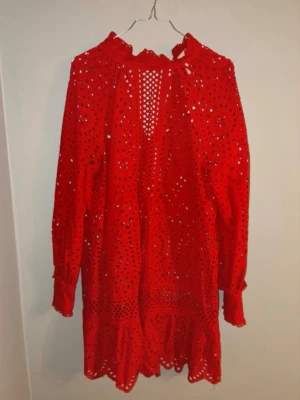 Röd broderad kortklänning med volang - Snygg röd kortklänning med broderade detaljer och volangkant. Klänningen har lång ärm, knappar vid ärmslut och en öppen detalj vid halsen med knyt. Perfekt för dig som vill sticka ut med en färgstark look.