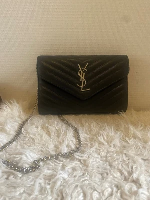 Svart YSL kuvertväska i läder - Snygg svart kuvertväska från YSL i quiltat läder med silverfärgad kedja och ikonisk YSL-logga framtill. Väskan har ett stilrent kuvertformat lock och elegant struktur. Perfekt för dig som vill ha en lyxig accessoar med tidlös design.