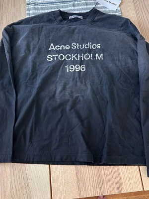 Svart långärmad topp Acne Studios - Svart långärmad topp från Acne Studios med tryck i vitt framtill där det står 'Acne Studios STOCKHOLM 1996'. Klassisk rund halsringning och relaxed fit. Perfekt till jeans eller kjol för en clean och stilren look.