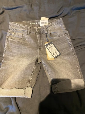 Gråa jeansshorts från Garcia - Snygga grå jeansshorts från Garcia med uppvikta benslut och klassisk femficksdesign. Modellen är slim fit med midwaist och har bälteshällor samt knapp- och dragkedjestängning. Perfekta för en avslappnad och trendig look.