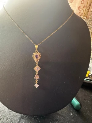 Guldigt halsband med lila stenar - Unikt halsband med tunn guldfärgad kedja och ett långt, hängande smycke med lila och klara stenar i olika former. Hänget har detaljerade utsmyckningar och flera geometriska former som ger en elegant och vintage-inspirerad look.