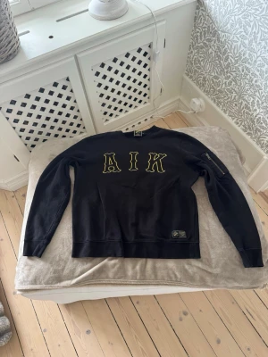 Svart AIK sweatshirt med gul text - Svart sweatshirt med stora gula bokstäver 'AIK' på bröstet. Tröjan har rund halsringning, lång ärm och en ficka med dragkedja på ena ärmen. Perfekt för dig som vill visa ditt AIK-support. Mjuk bomullskänsla. Går ej att få tag på längre.