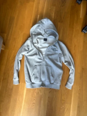 Grå hoodie från Peak Performance - Snygg grå hoodie från Peak Performance med dragkedja och broderad logga på bröstet. Hoodien har en stor ficka framtill och ribbade muddar vid ärmar och nederkant. Små fläckar på nedre ryggen! Perfekt för en avslappnad och sportig stil. OBS! Storlek 150 i barn!