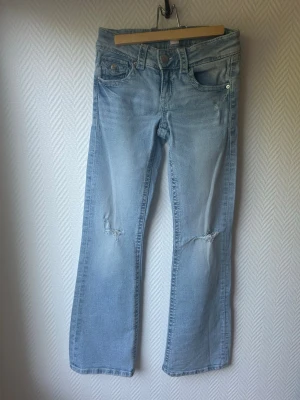 Ljusblå bootcut jeans med slitningar - Snygga ljusblå jeans med bootcut passform och slitningar vid knäna. Jeansen har klassiska fem fickor, knapp- och dragkedjegylf samt detaljerade bakfickor med lock. Perfekta för en avslappnad och trendig look.