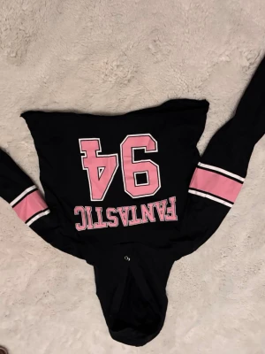 Hoodie - Svart hoodie med huva och trycket 'FANTASTIC 94' i rosa och vitt på framsidan. Ärmarna har breda rosa och vita ränder som ger en sportig vibe. Materialet känns mjukt och bekvämt.
