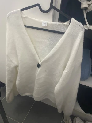 Vit fluffig kofta med dekorativ knapp - Mysig vit kofta i fluffigt material med djup V-ringning och en dekorativ svart knapp framtill. Koftan har lång ärm och avslappnad passform, perfekt att slänga på sig över en topp. Enkel och stilren design som passar till mycket.