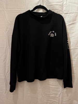 Svart sweatshirt med 90s-tryck - Cool svart sweatshirt med texten 'MADE IN THE 90s' tryckt på bröstet. Tröjan har en avslappnad passform, långa ärmar och ribbade muddar. Perfekt för dig som gillar retrostil och streetwear.den är lite nopprig 