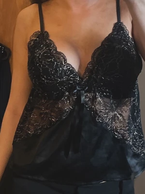 Svart spetstopp med satin och rosett - Snygg svart topp med tunna axelband, spetsdetaljer över bysten och genomskinliga partier. Nedre delen är i mjukt satinliknande material och framtill sitter en liten rosett. Perfekt för dig som gillar en elegant och sexig look.