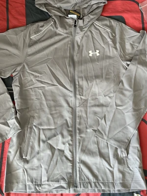 Grå vindjacka från Under Armour - Snygg grå vindjacka från Under Armour med huva och hel dragkedja framtill. Jackan har två sidofickor med dragkedja och en vit logga på bröstet. Tillverkad i lätt och slitstarkt material som passar perfekt för aktiva dagar utomhus.