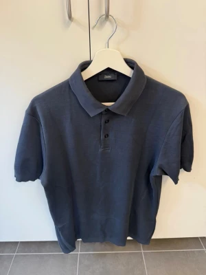 Mörkblå pikétröja från Herno, stl 50/M - Snygg mörkblå pikétröja från Herno med klassisk krage och knappar framtill. Tröjan har korta ärmar och är tillverkad i ett mjukt material som känns bekvämt mot huden. Perfekt för en stilren och avslappnad look.
