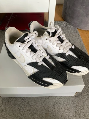 Nike vita och svarta sneakers - Säljer ett par unika Nike sneakers i vitt och svart med coola detaljer och kontrasterande paneler. Skorna har en låg silhuett, vita snören och en vit sula. Perfekta för dig som vill sticka ut med din stil.