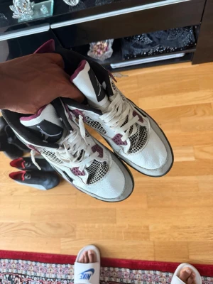 Vita och vinröda sneakers Air Jordan 4 - Snygga Air Jordan 4 sneakers i vitt läder med vinröda och svarta detaljer. Skorna har meshpaneler på sidorna, klassisk snörning och synlig Air-enhet i sulan. Perfekta för dig som gillar streetwear och vill sticka ut med en ikonisk modell.