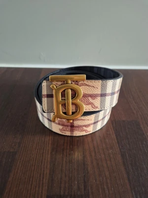 Burberry bälte - Snyggt bälte med klassiskt rutmönster i beige, brunt och rött samt ett stort guldigt B-spänne framtill. Bältet har svart insida och känns riktigt lyxigt. Perfekt accessoar för att lyfta din outfit.