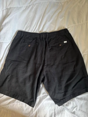 Svarta shorts från Selected Homme - Svarta shorts från Selected Homme med snörning i midjan och knappstängning. Shortsen har två bakfickor med knappar och är tillverkade i ett mjukt material som känns skönt mot huden. Perfekta för varma dagar.