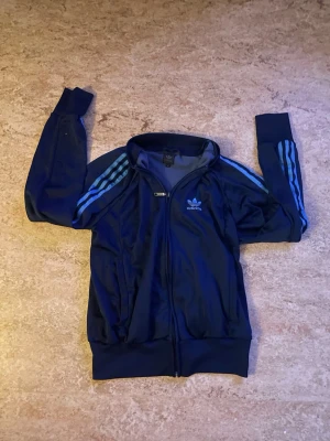 Adidas tröja - Snygg mörkblå tröja från Adidas med dragkedja framtill och klassiska ljusblå ränder längs ärmarna. Tröjan har ribbade muddar vid ärmslut och nederkant samt Adidas-logga på bröstet. Perfekt för sport eller chill. Storlek M, om ni vill ha mått är det bara att skriva!😊📏