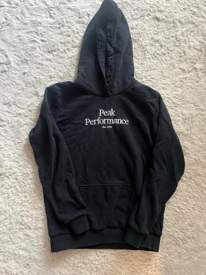 Svart hoodie från Peak Performance - Säljer en svart hoodie från Peak Performance med vit logga på bröstet. Klassisk modell med huva och känguruficka framtill. Mjuk insida och ribbade muddar vid ärmar och nederkant. Perfekt för chill dagar eller till skolan.