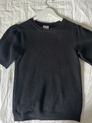 Svart stickad topp med puffärm - Snygg svart stickad topp från Vila med korta puffärmar och rund halsringning. Strukturen är småmönstrad och materialet känns mjukt och stretchigt. Perfekt till jeans eller kjol för en trendig look.